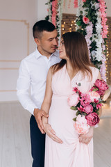 Charming young expecting couple poses in a white studio rich decorated with pink flowers