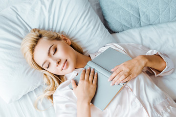 top view of young woman in pajamas holding book and sleeping on bed