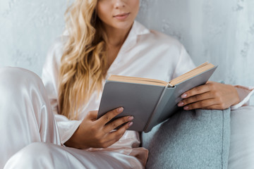 cropped shot of young woman in pajamas sitting on bed and reading book
