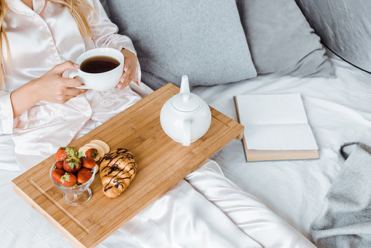 cropped image of woman in pajamas having breakfast in bed and holding cup of tea