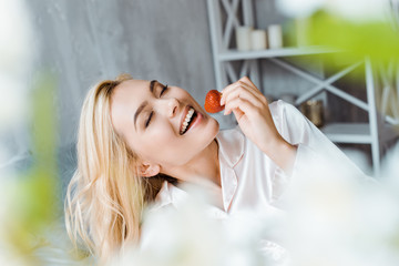 Obraz premium attractive woman in pajamas eating strawberry in bed in morning with closed eyes