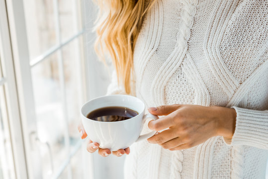 cropped image of woman in sweater holding cup of coffee near window at home