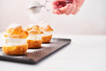 Sprinkling powdered sugar over delicious cream puff cakes on a black plate on white marble table over pink background. Selective focus, copy space.