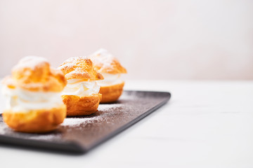 Delicious cream puff cakes with cream and powdered sugar on a black plate on white marble table over pink background. Selective focus, copy space.