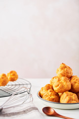 Bunch of fresh cream puff cakes on a white plate with ladle, whisk and wire rack on white marble table over pink background with copy space. Selective focus.