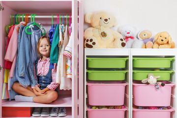 A little child girl is sitting in a wardrobe in a children's room. Saving and storage system for children's things and toys concept.