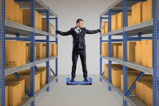 A businessman stands on a self-balancing scooter between two full storage racks.