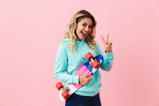Image of caucasian girl 20s smiling and holding skateboard, isolated over pink background