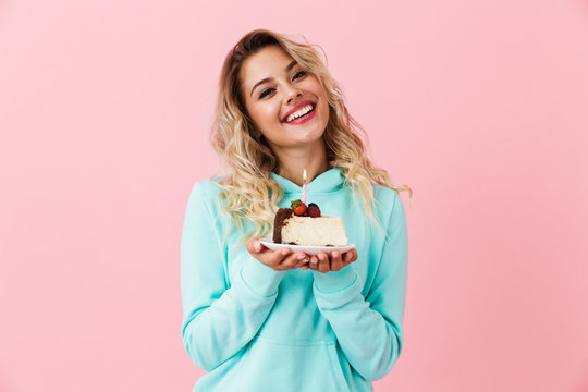 Photo of happy woman in basic clothing holding piece of birthday cake with candle, isolated over pink background