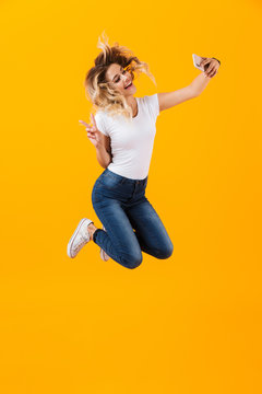 Full length image of young woman jumping and taking selfie photo on mobile phone, isolated over yellow background