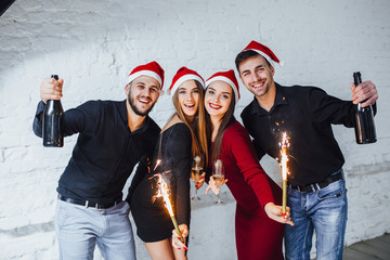 Four young friends hold champagne and fireworks in their hands, they are happy and celebrate the new year.