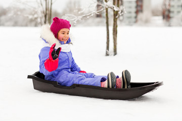 childhood, sledging and season concept - happy little girl in snowsuit sitting in sled outdoors in winter