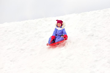 Fototapeta na wymiar childhood, sledging and season concept - happy little girl sliding down hill on snow saucer sled outdoors in winter