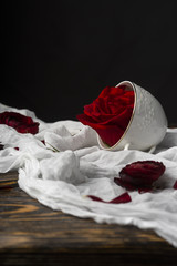 The buds of red roses, one of which is in a cup, lie on a crumpled white cloth on an old wooden table against a black background.