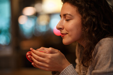 beautiful girl in gray knitted sweater sniffies fresh hot coffee from cup in restaurant. close up portrait. Autumn mood