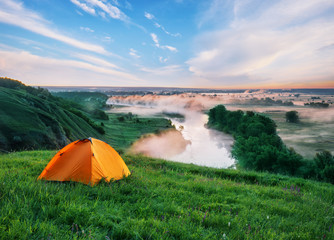 Orange tourist tent on a hill with green grass above the small r