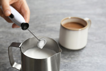 Woman using milk frother in pitcher near cup of coffee on table