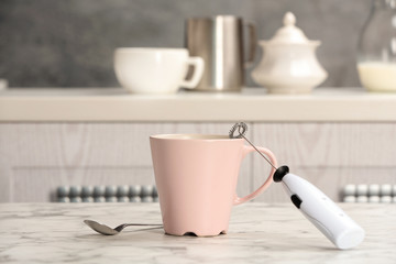 Milk frother device, cup of coffee and spoon on table in kitchen
