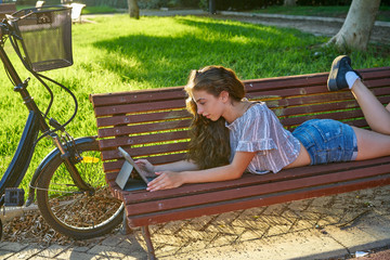 Girl lying on park bench playing with tablet