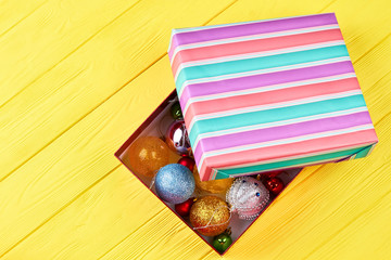 Gift box with Christmas decorations balls. Shiny Christmas baubles in multicolored striped box on yellow wooden background. Christmas holidays concept.