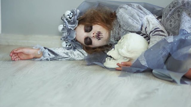 A dead dracula bride lies on the floor hugging a skull with one eye. Suddenly, she comes to life, opens her eyes. Child in halloween costume
