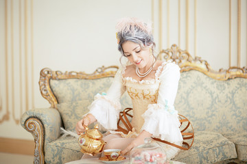 Beautiful greyhead woman in Rococo dress posing in front of pink background while pouring tea from the kettle