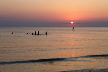 Fototapeta premium Lonely person paddling on paddle board and a group of kids are playing in the water during sunset