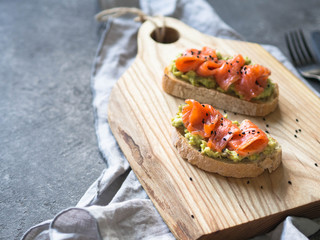 Toasts with avocado and salted salmon and black sesame seeds on a wood board on a gray background