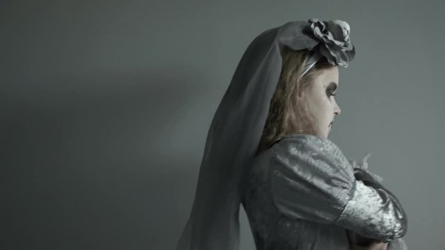 Teen girl in a costume of a dead bride with a face in makeup holding a witch's tumble in her hands. Passes by and leaves the frame with a sad face. Gray, cool colors