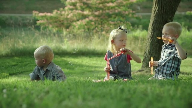 Happy three kids blowing soap bubbles in park seatting on the grass Slow motion Ful HD