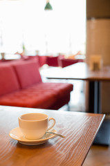 in the morning on a wooden table on the background of a red sofa and window is a cup of coffee or tea. there is a teaspoon on the saucer