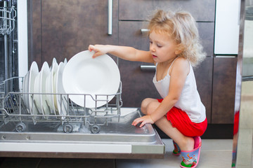 Cute blonde toddler girl helping in the kitchen taking plates out of dish washing machine