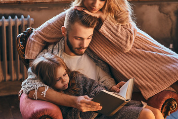 Family togetherness. Mom, dad and daughter reading story book together sitting on the couch. Family...