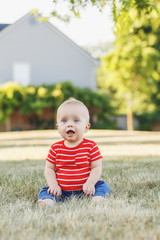 Cute adorable little Caucasian baby boy in red t-shirt and pants sitting in field meadow outside. Little happy smiling child in summer park on sunset. Lovely charming male infant having fun.