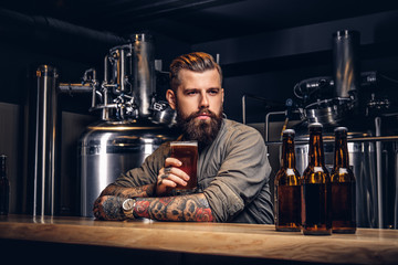 Tattooed hipster male with stylish beard and hair drinking beer sitting at the bar counter in the indie brewery.