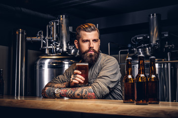 Tattooed hipster male with stylish beard and hair drinking beer sitting at the bar counter in the...