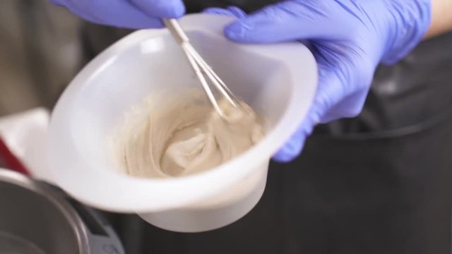 A close up of hands in blue rubber gloves holding a bowl with hair-dye and whisking it in circles, anticlockwise first, then clockwise