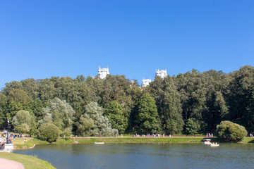 Fototapeta na wymiar Pond with green trees on the shore. The towers of the building in the blue sky, peeking from the trees. People boating on the lake and walk along the shore.