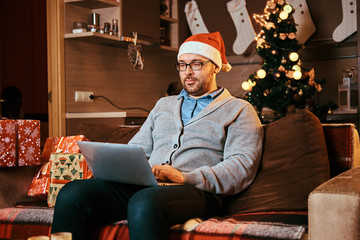 Man in Santa hat and glasses dressed in warm sweater working on laptop at Christmas.