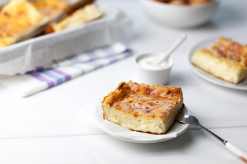 Tender, airy casserole of cottage cheese, is a piece on a plate, next to the baking sheet, sour cream in a gravy boat, standing on a table, on a light background, side view