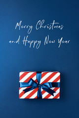 Christmas card with gift box wrapped in red striped paper and tied with blue bow on dark blue background. Holiday concept, top view.