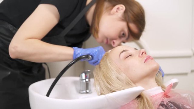 A close up of a hairdresser and a blonde holding her head in the backwash basin. The hairdresser has a shower and checks if the water is warm enough and starts washing the hair.