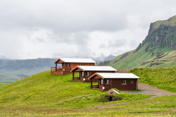 Three rustic wooden cabins in Vik, Iceland with cloudy, overcast grey stormy sky and cliff during summer, camping hotel accommodation