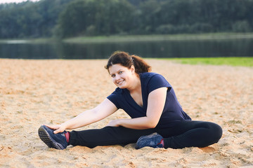 Fototapeta na wymiar Pretty woman working out and stretching legs while sitting on sand beach near lake.