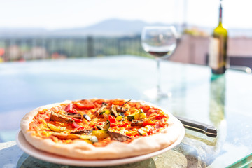 Macro closeup of fresh crust handmade pizza on glass table on terrace outside in Italy with red tomato sauce, vegetables, glass and bottle of red wine