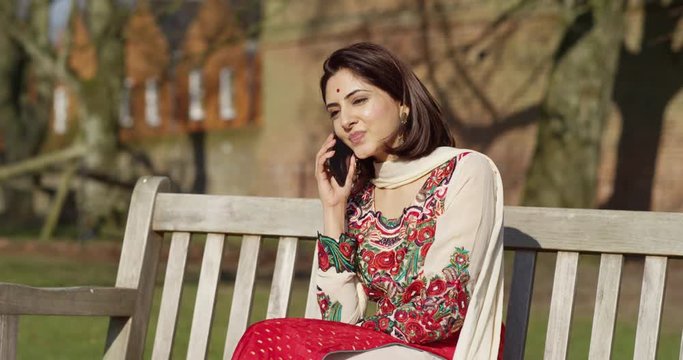 4K Beautiful young woman in traditional Indian dress talking on phone in the park. Slow motion.