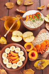Oatmeal with different fruits and honey. Oat flakes with milk and dried apricots on wooden background. Healthy food for vegan. Dry oats with berries of mountain ash and green mint. Autumn pattern