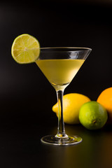 yellow cocktail with slice of lemon, and in the background a lemon, an orange and a lime on a black background