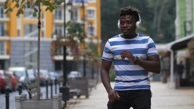 Portrait of joyful trendy afican american man in white headphones enjoying music and moving in afrohouse style dance on city street on rainy day. Groovy black guy dancing while listening music online.