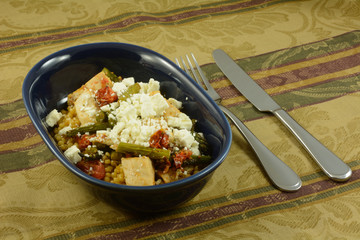 Israeli couscous or ptitim covered with chicken, roasted vegetables of asparagus and tomatoes with feta cheese on top in oval blue dish